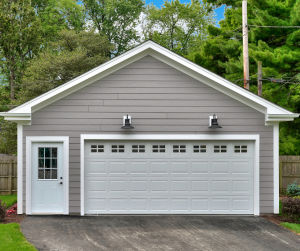 Two-car garage with a white paneled garage door and a single white service door to the right. There are two black sconce-style light fixtures above the garage door, and the building is painted in a light grey hue. The garage is set against a backdrop of greenery with a well-maintained asphalt driveway leading up to it.