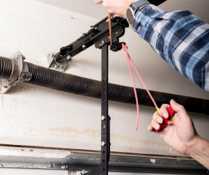 person's hands using tools to adjust or repair a garage door in San Diego. One hand is holding a red-handled pull cord (often an emergency release for garage doors), and the other hand is working on the track or spring system that assists in the door's operation.
