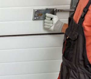 Person's hand wearing a white glove, turning a manual lock on a white garage door. The person is wearing an orange shirt and black overalls, suggesting they are a professional garage door technician or a worker engaged in maintenance or installation.