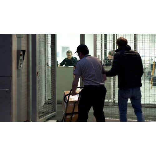 Security staff escorting a man with a luggage cart through a gated checkpoint at a secure facility.