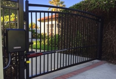 Black automatic swing gate with vertical iron bars, installed at the entrance of a residential driveway in Granite Hills, CA. The gate is equipped with a motorized opener and is framed by lush greenery and a stucco home with a red-tile roof in the background.