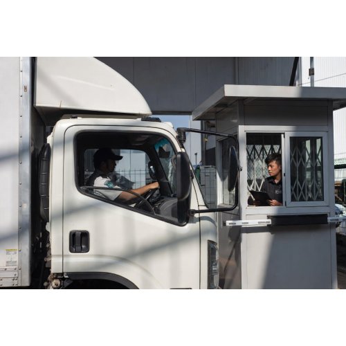 Security guard in a booth checking a delivery truck driver through a gated checkpoint.