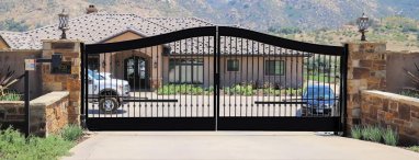 elegant black wrought iron swing gates at the entrance of a residential property near San Diego, CA. The gates have an arched top design and are mounted on substantial stone pillars. Each pillar is topped with a decorative lantern, adding to the aesthetic appeal of the entrance. Behind the gates, a driveway leads to a luxurious house with a tiled roof, surrounded by a well-manicured garden.