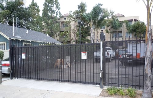 black sliding gate made of vertical metal bars, installed in a residential area near San Diego, CA. The gate is in a closed position, separating a parking lot from the street. Behind the gate, there are several parked vehicles, including a white pickup truck and a utility trailer.