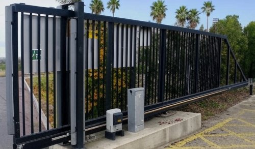 Black metal commercial slide gate with automated gate operator system installed at a business entrance, surrounded by palm trees and concrete curbing in Alpine, CA.