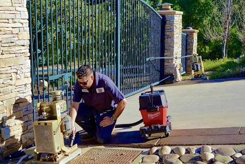 Technician is performing maintenance on a commercial gate opener system in San Diego, California. The gate is large, metal, and ornamental, with stone pillars on either side. The technician is kneeling on the ground, using tools to inspect or repair the gate mechanism. A toolbox and equipment are nearby, indicating a thorough maintenance process.