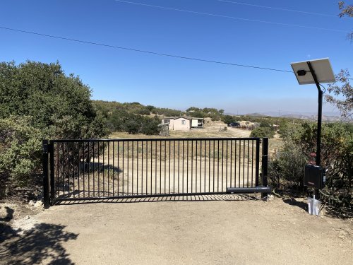 black metal sliding gate installed at the entrance of a rural property in Boulevard, California. The gate features vertical bars and is positioned across a dirt driveway. To the right of the gate, there is a solar panel mounted on a tall pole, likely powering the gate's automation system, with an associated control box below. 
