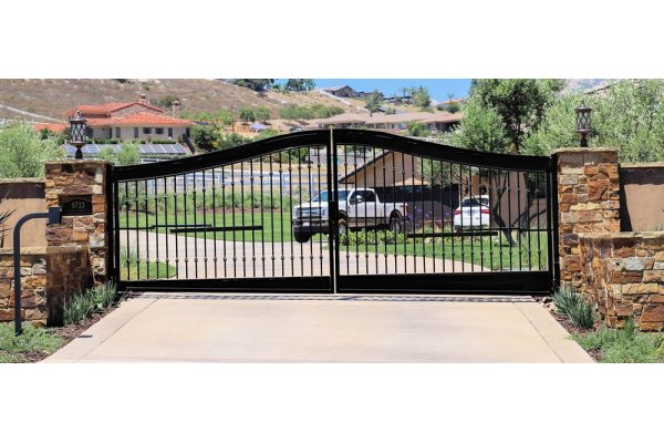 Black wrought iron gate at the entrance of a property in San Diego County California. The gate is designed with vertical bars and decorative scrolls at the top and center. It is attached to stone pillars on either side, which are capped with flat stone pieces. Beyond the gate, one can see a paved road with a white SUV parked on the opposite side. The background features a sunny suburban setting with manicured lawns, trees, and houses. 