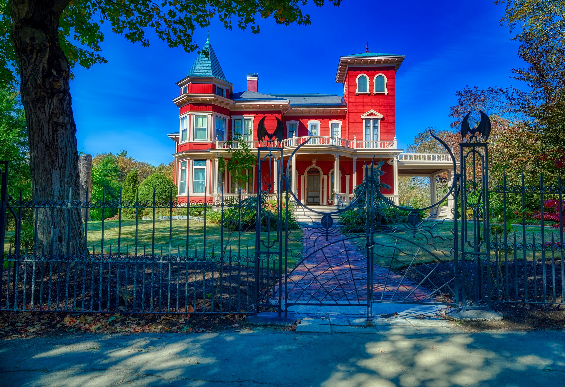 large, ornate Victorian-style house with red and blue exterior walls, a wraparound porch, and a distinctive turret. A decorative wrought iron fence with an open gate stands in the foreground, and the house is surrounded by a well-kept lawn with trees, under a clear blue sky.