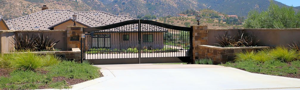 Black wrought iron driveway gate with stone pillars and surrounding desert landscaping in San Diego County, California.
